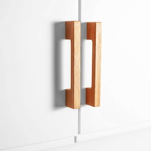 Könnte beinhalten: Zwei hölzerne Schrankgriffe mit hellbraunem Finish sind an einer weißen Schranktür montiert. Die Griffe haben eine rechteckige Form mit einem Ausschnitt in der Mitte, wodurch ein Griff entsteht.