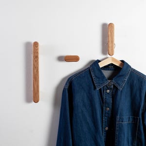 Könnte beinhalten: Drei Wandhaken aus Holz in minimalistischem Design. Zwei sind vertikal, länglich, einer horizontal und oval. Ein Jeanshemd hängt an einem Holzkleiderbügel an einem der vertikalen Haken. Die Haken sind an einer weißen Wand befestigt.