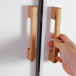 Könnte beinhalten: Zwei Holzschrankgriffe mit hellbraunem Finish sind an einer weißen Schranktür montiert. Ein Griff wird von einer Hand gehalten. Die Griffe haben ein modernes, minimalistisches Design.