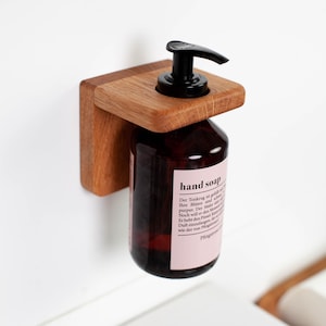 Könnte beinhalten: Ein wandmontierter Holzhalter für eine dunkel bernsteinfarbene Glasflasche mit schwarzem Pumpenspender. Die Flasche hat ein hellrosa Etikett mit dem Text hand soap und deutschem Text. Die Holzhalterung weist eine natürliche Maserung auf.