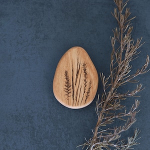 Puede incluir: Una pieza decorativa de madera en forma de lágrima con tallos de trigo grabados. La madera de color claro contrasta con el fondo oscuro y la ramita seca. El diseño de trigo añade un elemento natural y rústico.