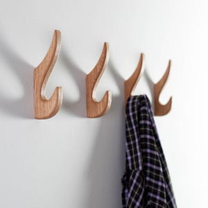 May include: Four wooden wall hooks with a curved design, mounted on a white wall. The hooks are made of light-colored wood and have a smooth finish. A black and purple plaid shirt hangs on one of the hooks.