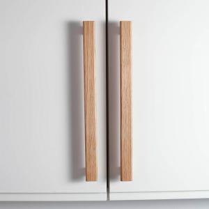 Könnte beinhalten: Zwei hellfarbene Holzschrankgriffe sind vertikal an einer weißen Schranktür angebracht. Die Griffe sind rechteckig und haben eine natürliche Holzmaserung. Die Schranktür ist geschlossen, mit einer dünnen schwarzen Linie zwischen den beiden Türen.