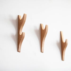 Könnte beinhalten: Drei Wandhaken aus Holz mit natürlicher Oberfläche sind an einer weißen Wand befestigt. Die Haken haben ein geschwungenes Design, mit einem Doppel-Haken am größten. Die Holzmaserung ist sichtbar und verleiht dem minimalistischen Design Textur.