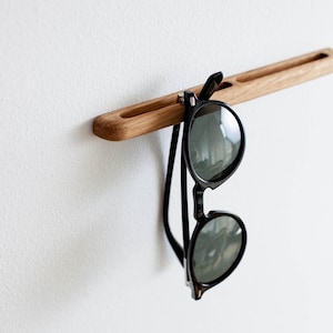 May include: A wooden wall-mounted sunglasses holder with two slots. A pair of black sunglasses is hanging from the holder.