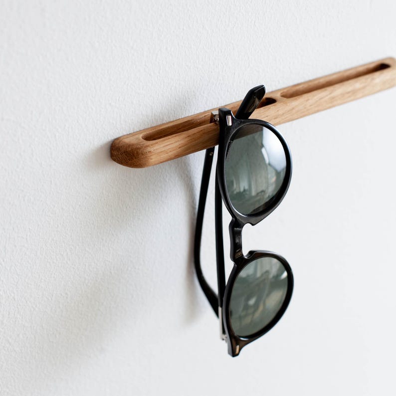 May include: A wooden wall-mounted sunglasses holder with two slots. A pair of black sunglasses is hanging from the holder.