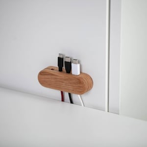 May include: A wooden cable organizer with three charging cables. The organizer is made of light brown wood and has a curved shape. The cables are black and white.