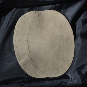 May include: Two beige suede leather patches with a stitched edge. The patches are oval shaped and are lying on a black fabric surface.