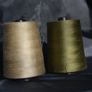 May include: Two spools of thread, one beige and one olive green, are sitting on a dark surface.
