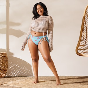 May include: A light blue bikini bottom with a flamingo pattern. The bikini bottom has turquoise ties on each side. The model is wearing a beige crochet top and a white bikini top. The background is a light beige color.