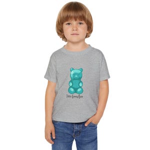 May include: A gray t-shirt featuring a teal gummy bear graphic with the text "Little Gummy Bear" in a script font. The shirt is worn by a young child wearing blue jeans.