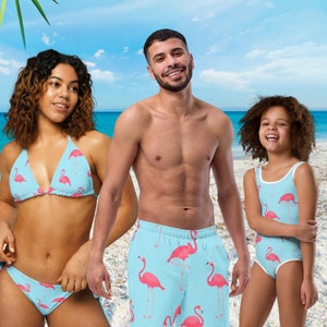 以下が含まれることがあります： フラミンゴ柄のマッチングスイムウェアを着た3人。スイムウェアはピンクのフラミンゴが描かれたブルー。人々はビーチに立っています。