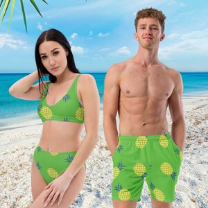 May include: A green bikini and matching swim trunks with a yellow pineapple pattern. The bikini top has a scoop neck, and the high-waisted bottoms and swim trunks have a relaxed fit. The swimwear is set against a beach backdrop.