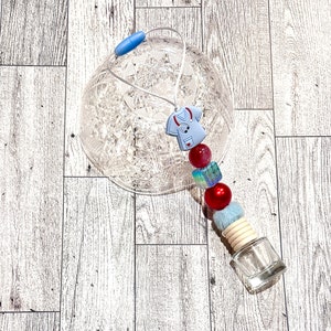 Puede incluir: Un amuleto de bata de médico azul y blanco con cuentas rojas y azules, un pompón blanco esponjoso y una botella de vidrio transparente con una tapa de madera. El amuleto cuelga de un ambientador de coche de plástico transparente.