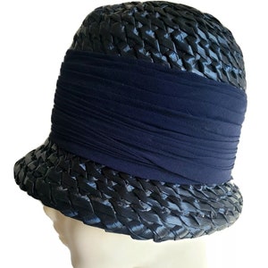 May include: A navy blue cloche hat with a woven straw design. The hat features a wide, pleated navy band around the crown. The brim is slightly curved, and the hat is displayed on a mannequin head.