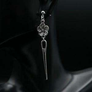 May include: Silver dangle earrings with a flower charm and a needle-shaped pendant.