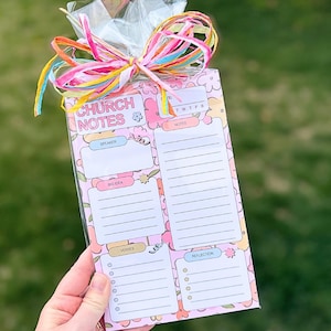 May include: A pink floral Church Notes notepad with sections for speaker, big ideas, verses, and reflection. The notepad is wrapped in clear cellophane and tied with a colorful ribbon. The text "CHURCH NOTES" is at the top.