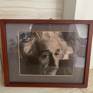 May include: A framed black and white photograph of Albert Einstein. The photo shows Einstein's face and upper torso. He is looking to the right of the frame. The photo is framed in a dark wood frame with a mat.