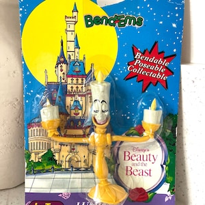 May include: A yellow plastic bendable figurine of Lumiere, the candelabra from Disney's Beauty and the Beast. The figurine is posed with its arms outstretched, holding two candles. The packaging features a cartoon image of the Beast's castle and the text "Bend-Ems Bendable Poseable Collectable".
