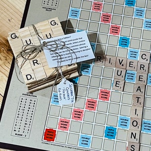 May include: A set of wooden Scrabble tile coasters with the words "Scrabble Tile Coasters - Rebus Edition". The tiles are arranged to spell the word "GUILD". The coasters are on a blue and white Scrabble game board.