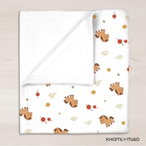 May include: White baby blanket with a repeating pattern of cartoon horses, red lanterns, yellow flowers, and clouds. The blanket is folded over, revealing a plain white interior. The blanket has a soft, cozy appearance.