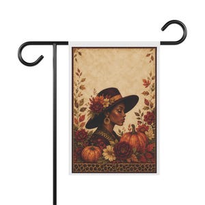 Puede incluir: Una bandera de jardín rectangular con una ilustración de perfil de una persona con un sombrero de ala ancha adornado con flores de otoño. La escena incluye calabazas naranjas, follaje otoñal rojo y amarillo y un borde de leopardo.