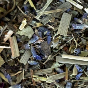 Puede incluir: Primer plano de una mezcla de hierbas y flores secas, incluyendo lavanda, hierba de limón y otras hierbas verdes. La mezcla es de color marrón claro con toques de azul y verde.