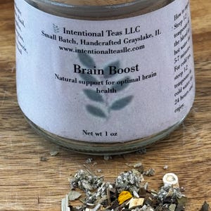 May include: A glass jar with a bamboo lid containing Brain Boost tea blend from Intentional Teas LLC. The label reads "Brain Boost" and "Natural support for optimal brain health". The jar is sitting on a wooden surface with a small pile of the tea blend scattered in front of it.