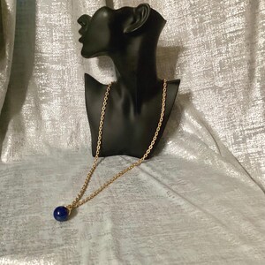May include: A gold-colored chain necklace with a dark blue, round pendant. The necklace is displayed on a black bust against a silver, textured background. The pendant is a deep, solid blue color.