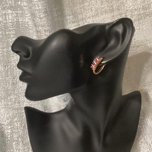 May include: A gold hoop earring featuring a row of small, vibrant red gemstones set along the outer edge. The earring is displayed on a black mannequin head, showcasing the curved design and the contrast between the gold metal and red stones.