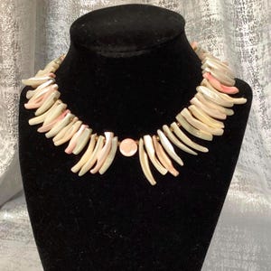 May include: A shell necklace displayed on a black mannequin. The necklace features cream-colored, elongated shell pieces with pink accents. A round, pink shell is centered on the necklace, with gold-colored beads connecting the shells.