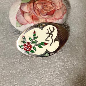 May include: Oval-shaped belt buckle with a cream-colored background, featuring a red rose and a deer silhouette. The buckle has a decorative border and is set against a white surface. A painted rock with a rose design is in the background.