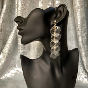 May include: Clear, dangling earrings with a gold-colored stud. The earrings are made of multiple, circular, transparent pieces that cascade down. The earrings are displayed on a black mannequin head against a silver, textured backdrop.