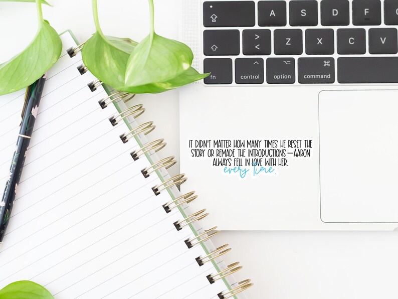 May include: A white spiral notebook with lined pages, a black pen with a floral design, and a laptop keyboard. The image includes a sticker with the text "IT DIDN'T MATTER HOW MANY TIMES HE RESET THE STORY OR REMADE THE INTRODUCTIONS - AARON ALWAYS FELL IN LOVE WITH HER. every time."