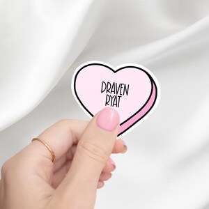 May include: A pink heart-shaped sticker with the words "DRAVEN RYAT" in black, held by a hand with a gold ring and pink nail polish. The sticker has a black outline and a pink interior. The background is a white, silky fabric.