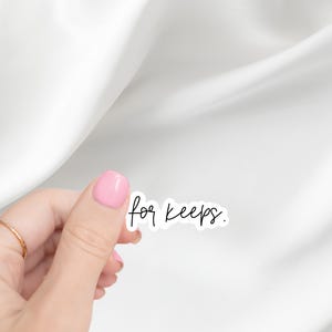 May include: A white sticker with the phrase "for keeps" in a handwritten black font. The sticker is held by a hand with pink nail polish and a gold ring, set against a white background.
