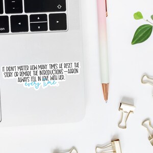 May include: A white sticker with black text that reads: "IT DIDN'T MATTER HOW MANY TIMES HE RESET THE STORY OR REMADE THE INTRODUCTIONS - AARON ALWAYS FELL IN LOVE WITH HER. every time." A pink and white pen, gold binder clips, and a laptop are also visible.