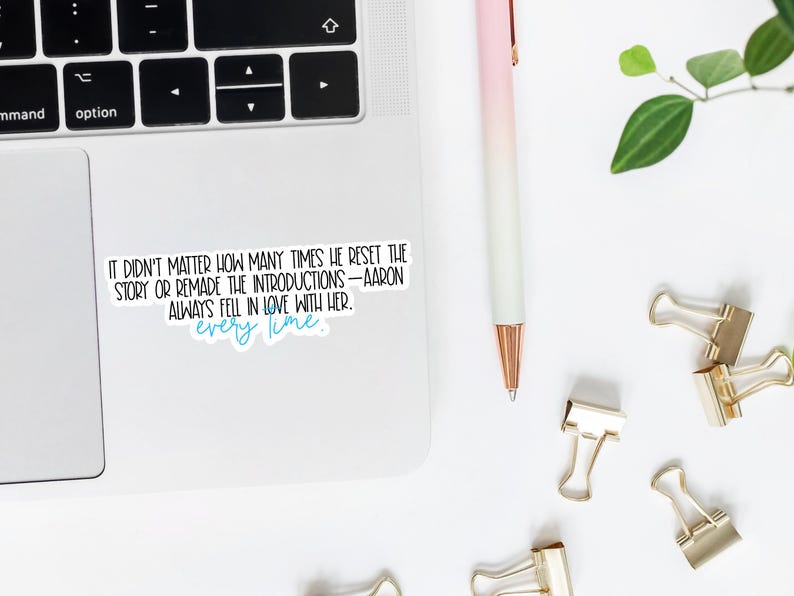 May include: A white sticker with black text that reads: "IT DIDN'T MATTER HOW MANY TIMES HE RESET THE STORY OR REMADE THE INTRODUCTIONS - AARON ALWAYS FELL IN LOVE WITH HER. every time." A pink and white pen, gold binder clips, and a laptop are also visible.
