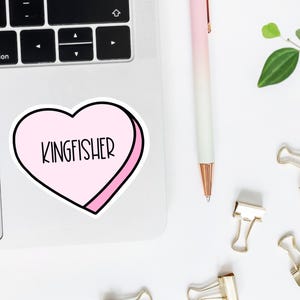 K&ouml;nnte beinhalten: Ein rosa herzf&ouml;rmiger Aufkleber mit wei&szlig;em Rand und dem Wort "KINGFISHER" in schwarzer Schrift darin. Der Aufkleber befindet sich auf einem silbernen Laptop.