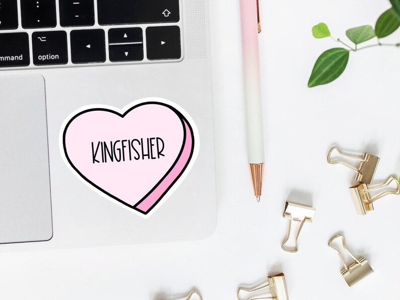 K&ouml;nnte beinhalten: Ein rosa herzf&ouml;rmiger Aufkleber mit wei&szlig;em Rand und dem Wort "KINGFISHER" in schwarzer Schrift darin. Der Aufkleber befindet sich auf einem silbernen Laptop.