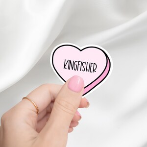 K&ouml;nnte beinhalten: Ein rosa herzf&ouml;rmiger Aufkleber mit wei&szlig;em Rand und dem Wort "KINGFISHER" in schwarzer Schrift darin.