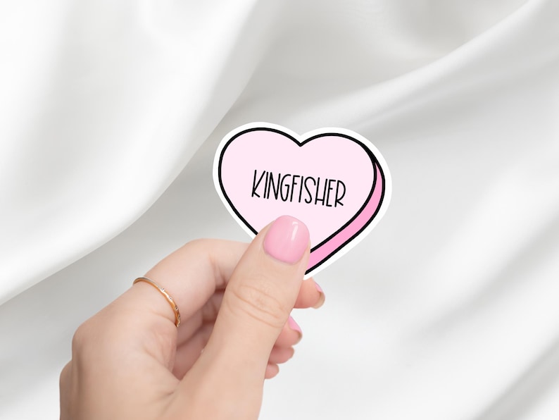 K&ouml;nnte beinhalten: Ein rosa herzf&ouml;rmiger Aufkleber mit wei&szlig;em Rand und dem Wort "KINGFISHER" in schwarzer Schrift darin.