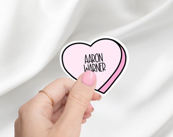 May include: A pink heart-shaped sticker with a white outline and the text "AARON WARNER" inside.