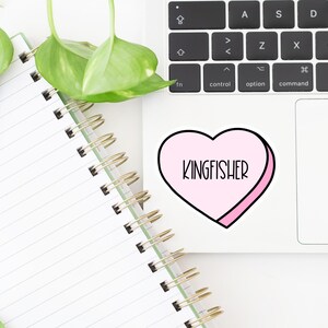 K&ouml;nnte beinhalten: Ein rosa herzf&ouml;rmiger Aufkleber mit wei&szlig;em Rand und dem Text "KINGFISHER" in schwarzen Buchstaben.