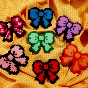 Op de afbeelding: Een verzameling gepixelde strikvormige knutselwerkjes in verschillende kleuren, waaronder roze, blauw, groen, paars en oranje, elk omlijnd in zwart. De strikken zijn gerangschikt op een gele, getextureerde ondergrond.