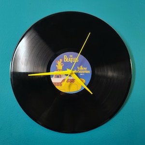 Peut inclure: Une horloge en vinyle noir avec l'illustration "Yellow Submarine" des Beatles. L'horloge a des aiguilles jaunes et une étiquette bleue et jaune. L'horloge est fixée sur un mur turquoise.