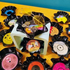 May include: Several black vinyl record bowls, some with items like sunglasses and keys. The bowls are displayed on a yellow patterned surface. Some bowls feature record labels with text and colourful designs.