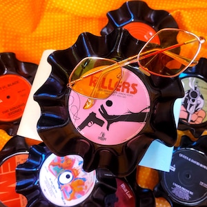 May include: A pair of gold-rimmed sunglasses with orange-tinted lenses resting on a black vinyl record bowl. The record label features the text The Killers, Mr. Brightside, and a graphic of a hand and a handgun. Other records are visible nearby.