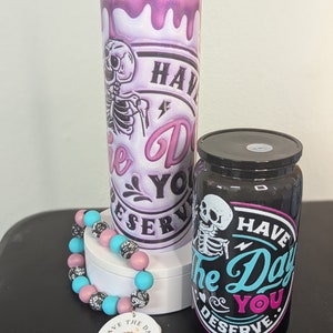 Könnte beinhalten: Zwei Becher mit einem Skelett-Motiv und dem Text "Have the Day You Deserve". Ein Becher ist rosa und der andere ist schwarz. Ein rosa-blaues Perlenarmband mit einem Anhänger, auf dem "Have the Day You Deserve" steht, befindet sich im Vordergrund.