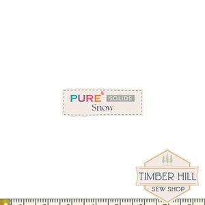 May include: A white fabric label with the text "PURE SOLIDS Snow" in teal, orange, and purple. The label is rectangular with a dotted border. Below the label is a measuring tape showing inches. A Timber Hill Sew Shop logo is also visible.